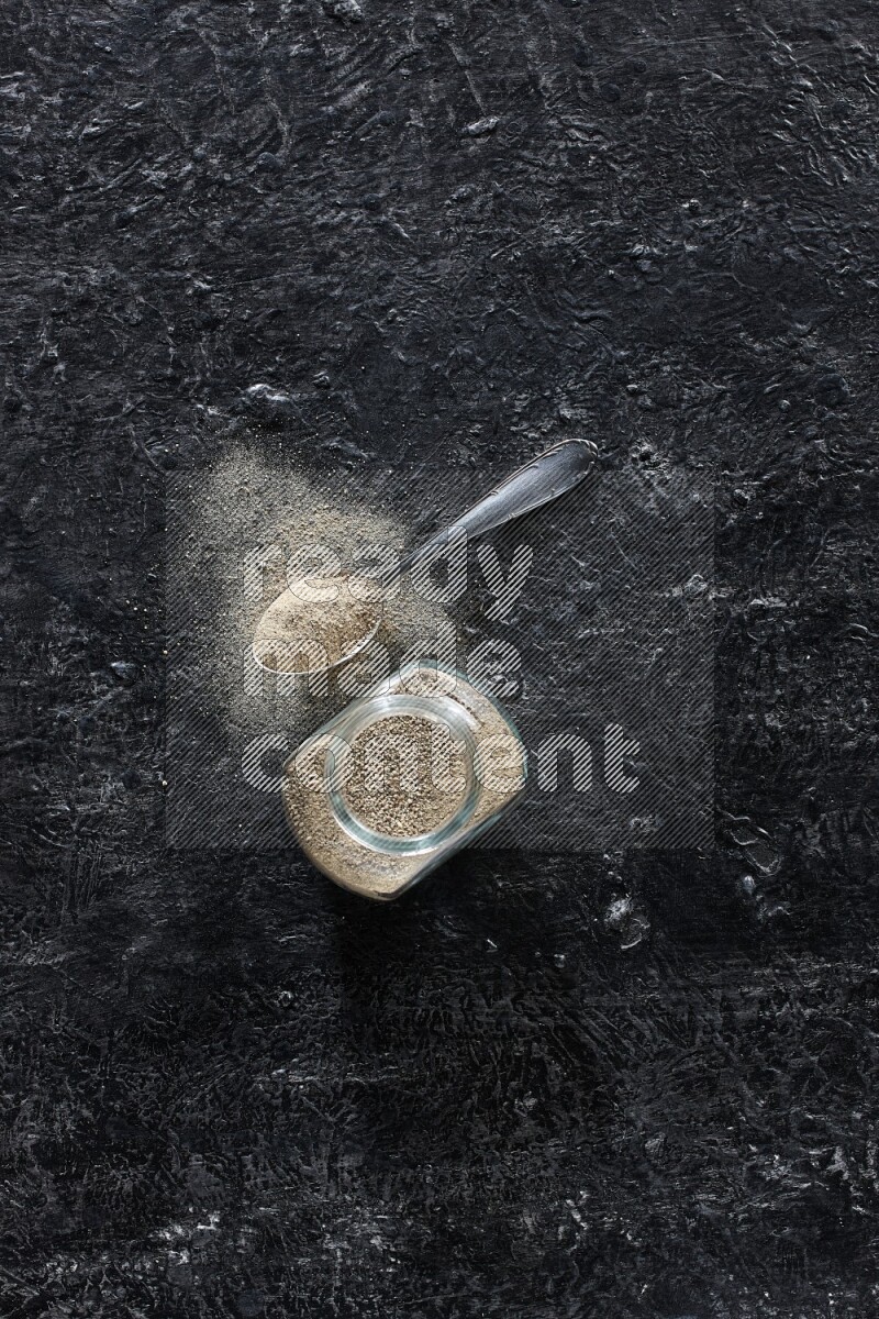 وعاء زجاجي وملعقة معدنية مملوءة بمسحوق الفلفل الأبيض على أرضية سوداء