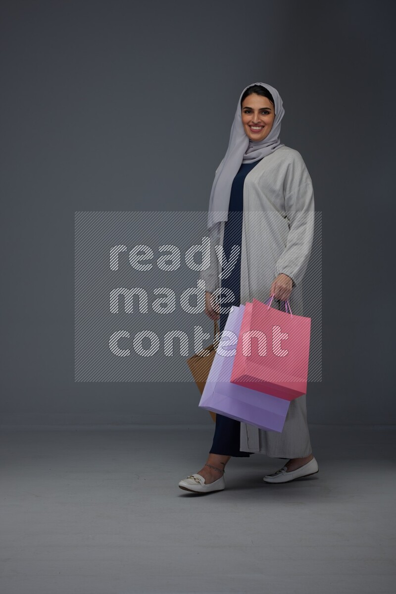 A Saudi woman wearing a light gray Abaya and head scarf standing and holding shopping bags on a grey background
