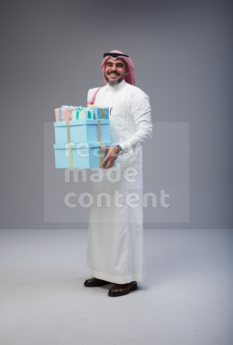 Saudi man Wearing Thob and red Shomag standing holding gift box on Gray background