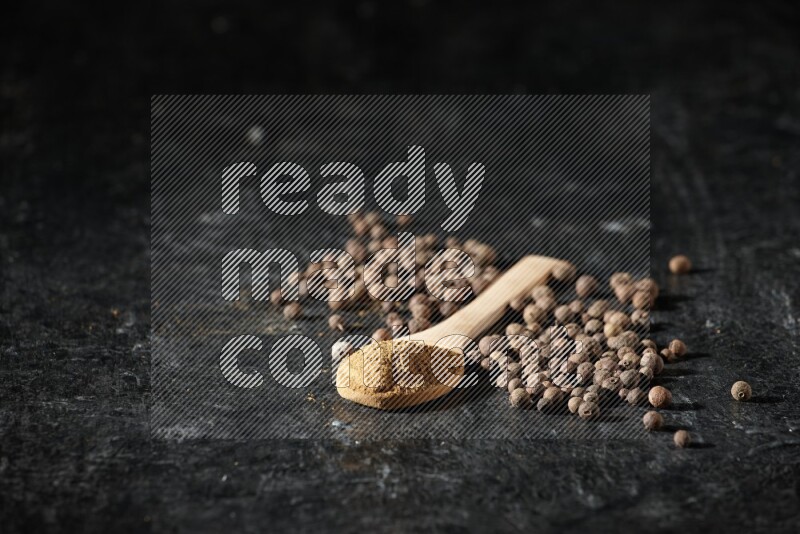 Wooden spoons full of all spice powder and allspice whole balls beside it on a textured black flooring