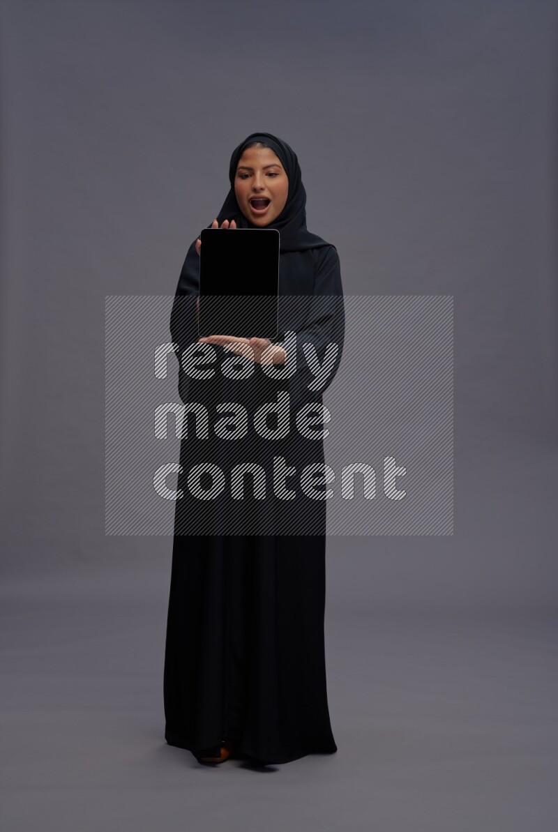 Saudi woman wearing Abaya standing showing tablet to camera on gray background