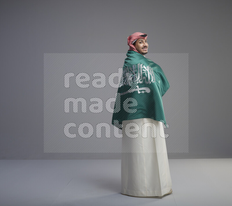 A Saudi man standing wearing thob and red shomag wrapping big saudi flag on gray background