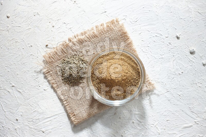 A glass bowl full of cumin powder with some of cumin seeds on burlap piece on a textured white flooring