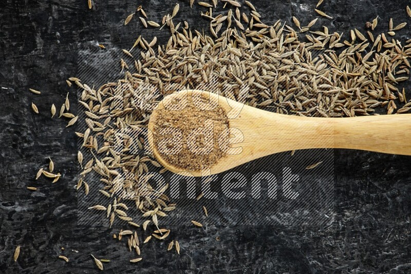 A wooden spoon full of cumin powder and cumin seeds spreaded on textured black flooring