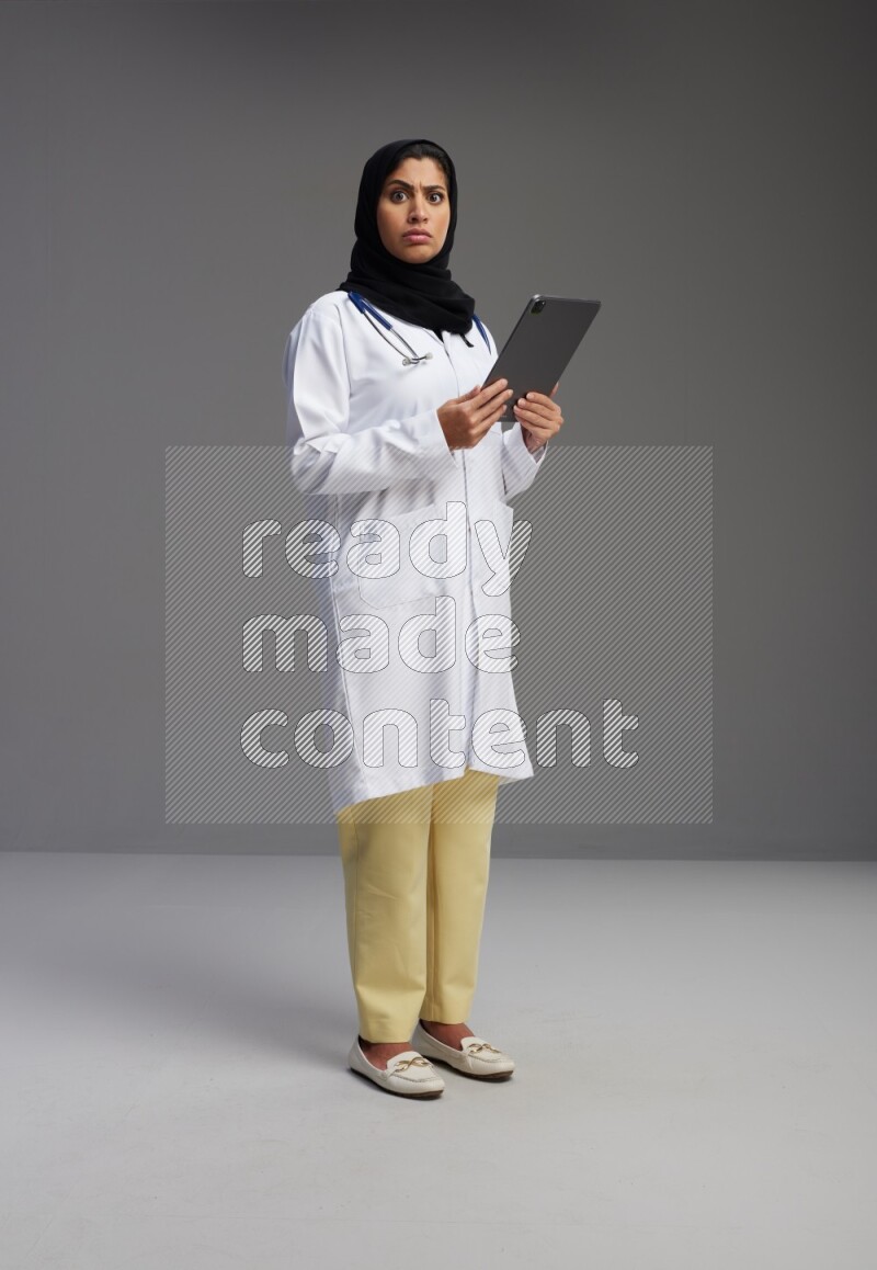 Saudi woman wearing lab coat with stethoscope standing working on tablet on Gray background