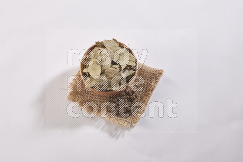A wooden bowl filled with dried bay leaves on a piece of burlap with some of allspice berries on white flooring in different angles