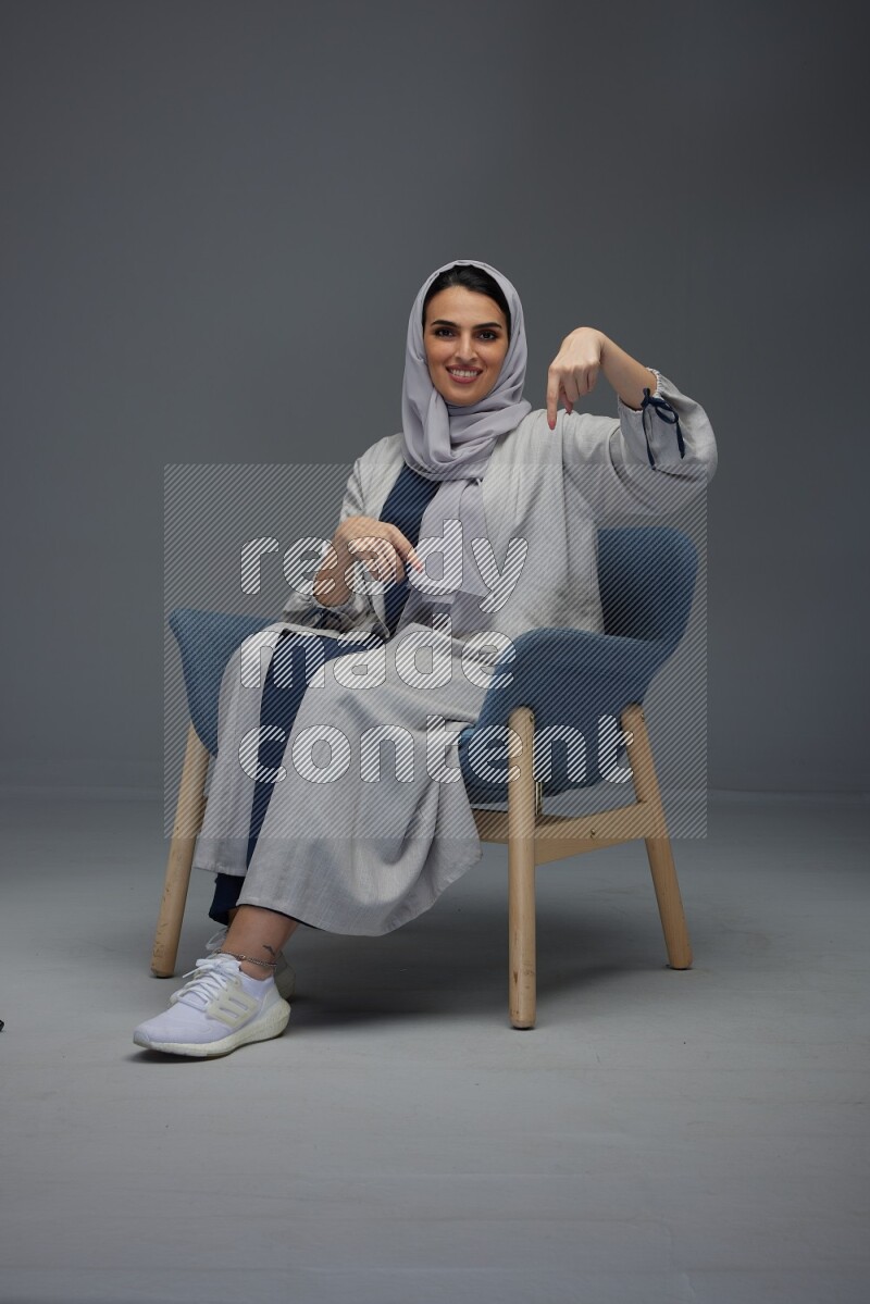 A Saudi woman wearing a light gray Abaya and head scarf sitting on a dark grey chair while making multi hand gestures eye level on a grey background