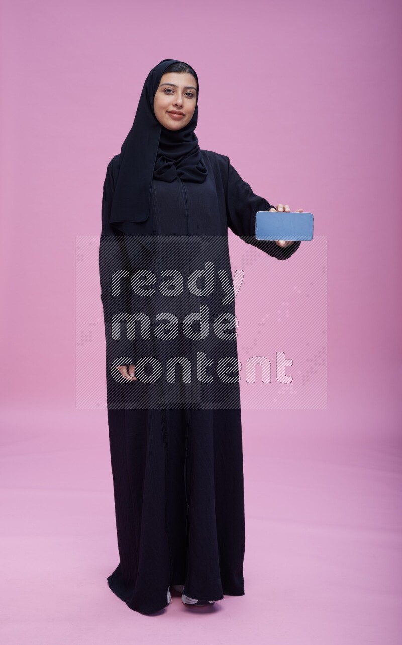 Saudi woman wearing Abaya standing showing phone to camera on pink background
