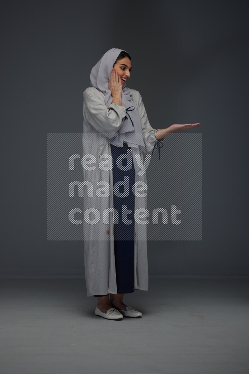 A Saudi woman wearing a light gray Abaya and head scarf standing and pointing in different directions eye level on a grey background