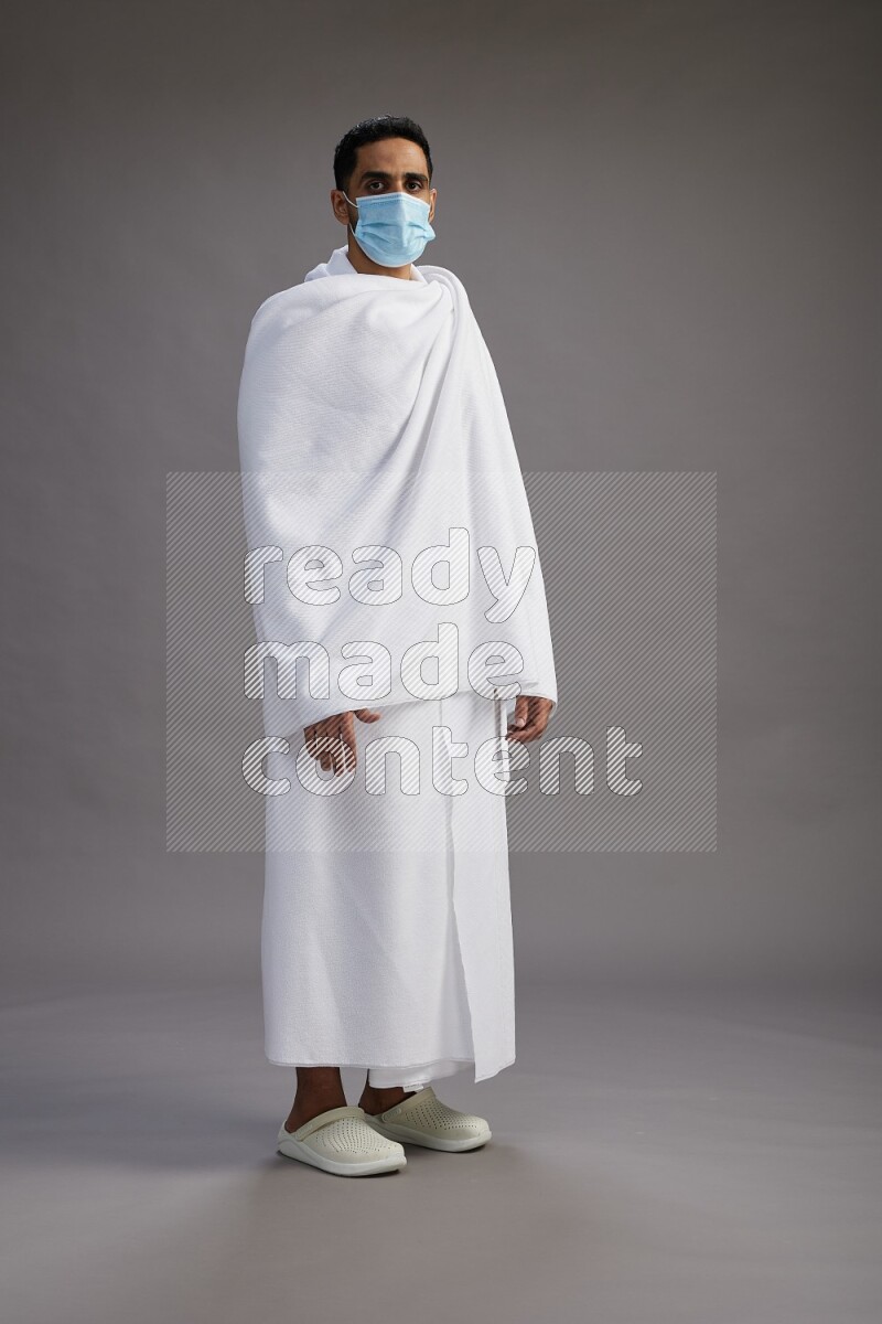 A man wearing Ehram with face mask Standing Interacting with the camera on gray background