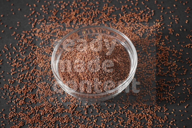 A glass bowl full of garden cress seeds surrounded by seeds on a black flooring