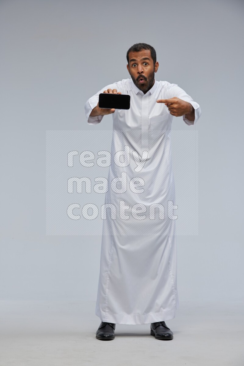 Saudi man wearing Thob standing showing phone to camera on Gray background