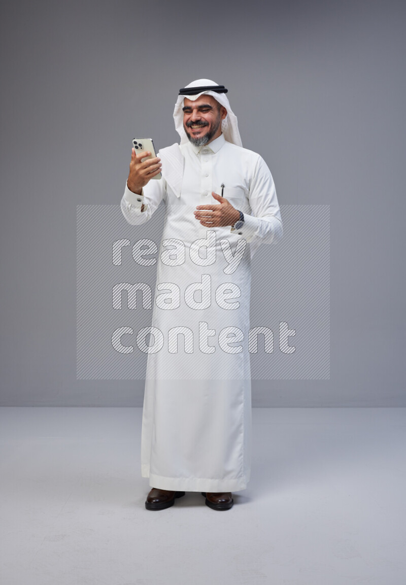 Saudi man Wearing Thob and white Shomag standing texting on phone on Gray background