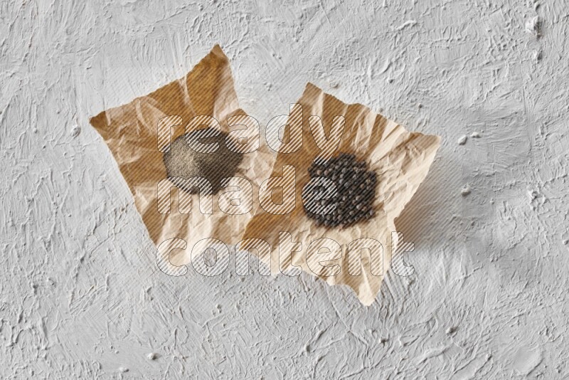 Black pepper and black pepper powder on 2 crumpled pieces of craft paper on a textured white flooring