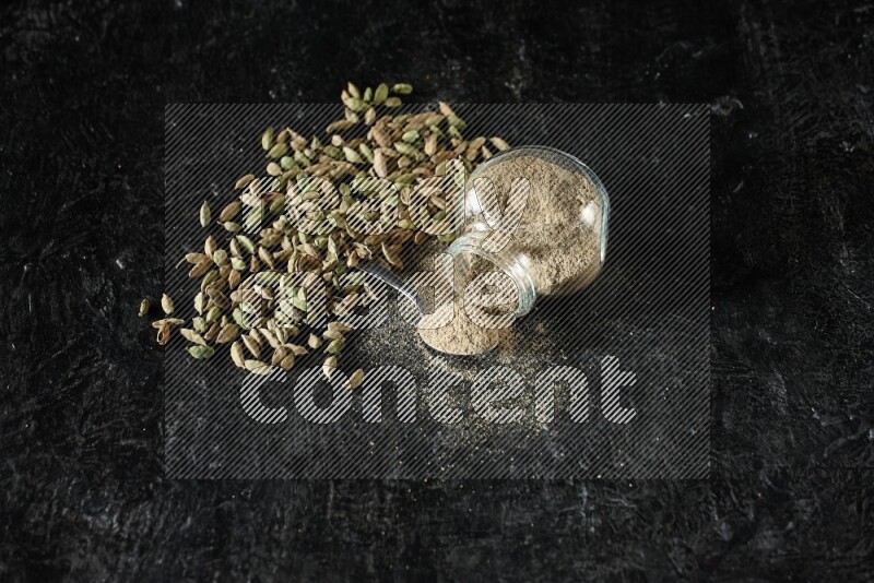 A flipped glass spice jar and a metal spoon full of cardamom powder and cardamom seeds spreaded on textured black flooring