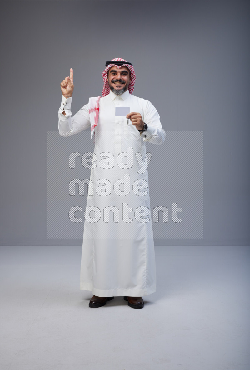 Saudi man Wearing Thob and red Shomag standing holding ATM card on Gray background