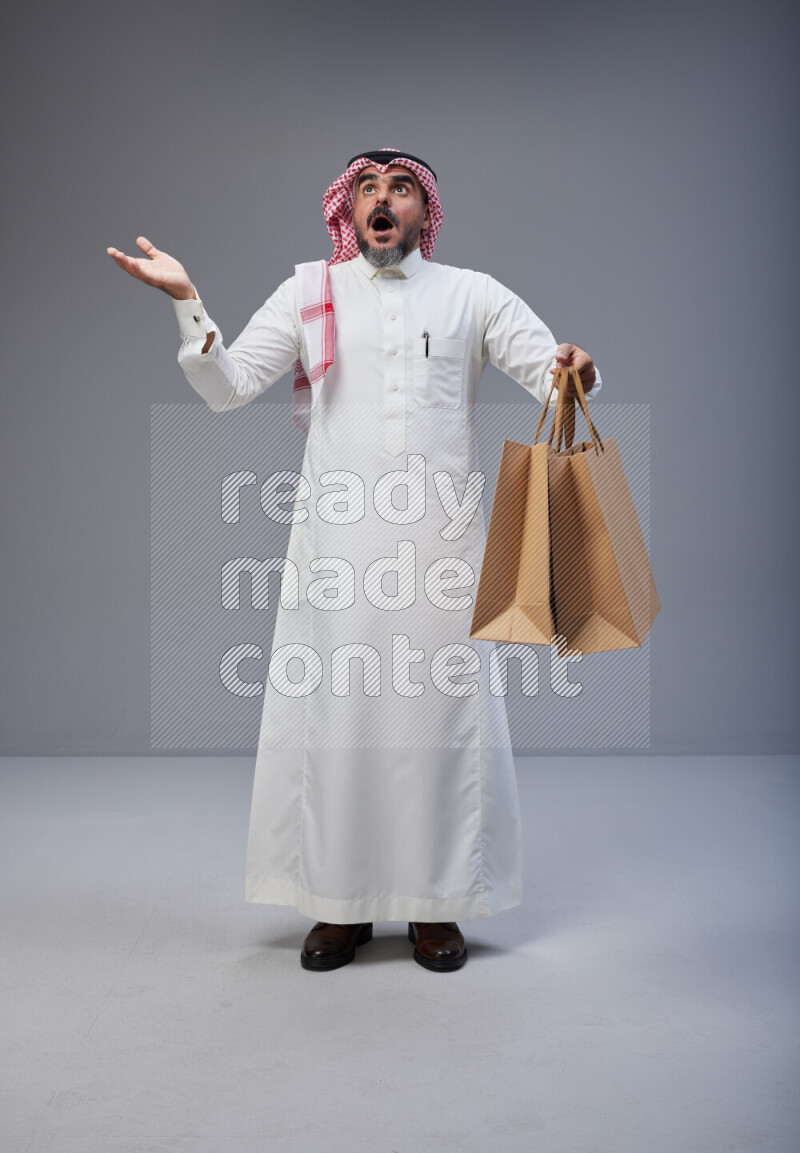 Saudi man Wearing Thob and red Shomag standing holding shopping bag on Gray background