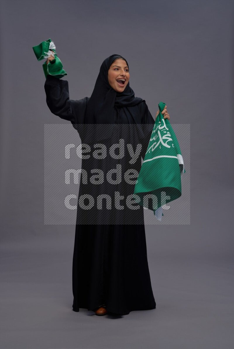 Saudi woman wearing Abaya standing holding Saudi flag on gray background