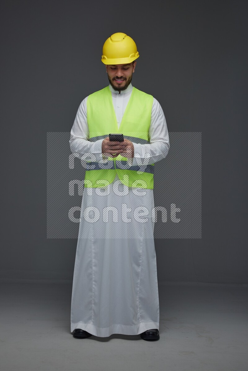 A Saudi man wearing Thobe with a yellow safety vest and white helmet standing and using his phone eye level on a gray background