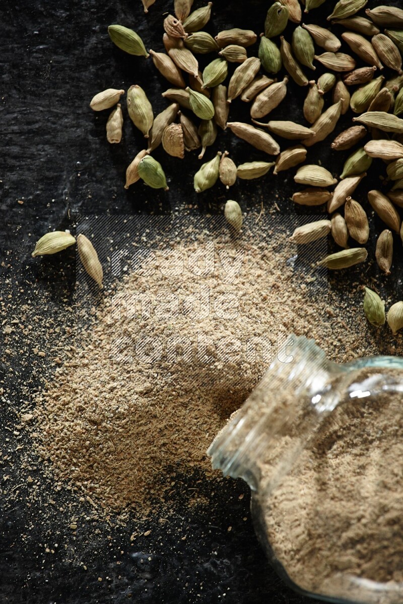 وعاء زجاجي للتوابل مقلوب وممتلئ ببودرة الهيل والبودرة تناثرت منه مع حبوب الهيل المتناثرة على خلفية سوداء