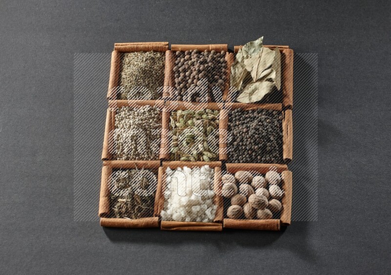 9 مربعات من أعواد القرفة ممتلئة بالهيل في المنتصف محاطة بجوزة الطيب والفلفل الأسود وورق الغار والبهارات والكمون والفلفل الأبيض والريحان المجفف والملح على أرضية سوداء