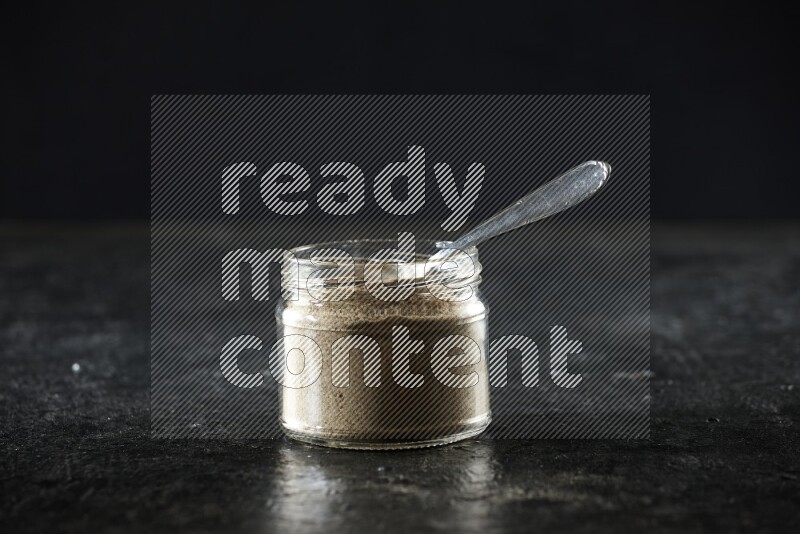 A glass jar and metal spoon full of cardamom powder on textured black flooring