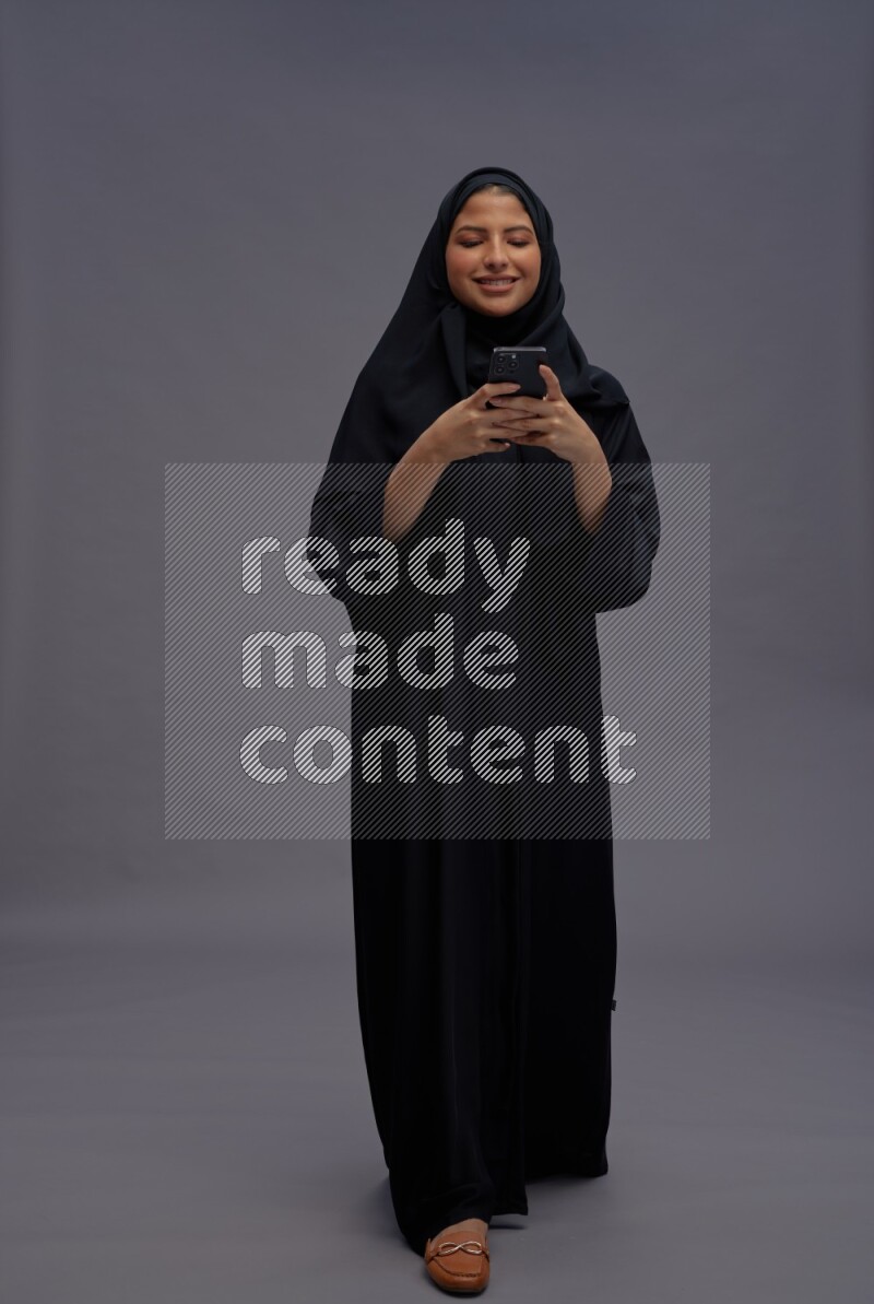 Saudi woman wearing Abaya standing texting on phone on gray background
