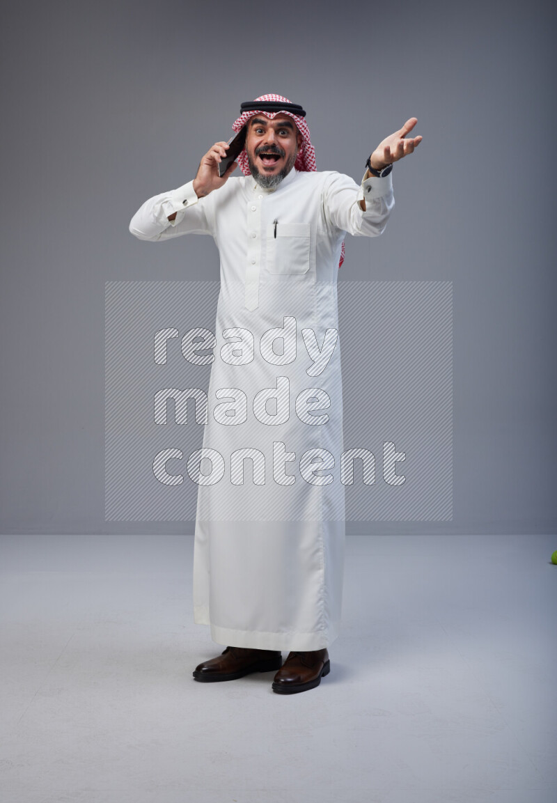 Saudi man Wearing Thob and red Shomag standing talking on phone on Gray background
