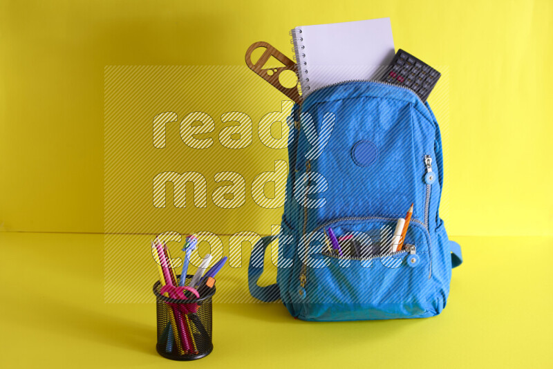A school bag with assorted school supplies in and beside it on yellow background
