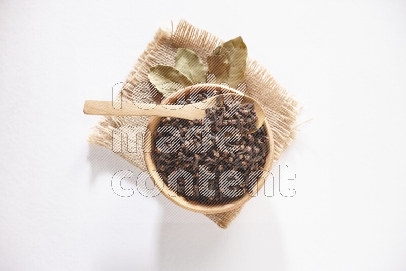 A wooden bowl and a wooden spoon full of cloves on a piece of burlap on a white flooring