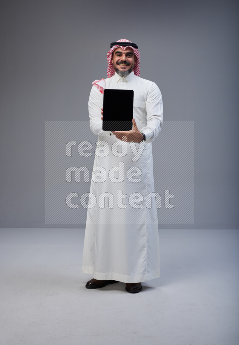 Saudi man Wearing Thob and red Shomag standing showing tablet to camera on Gray background