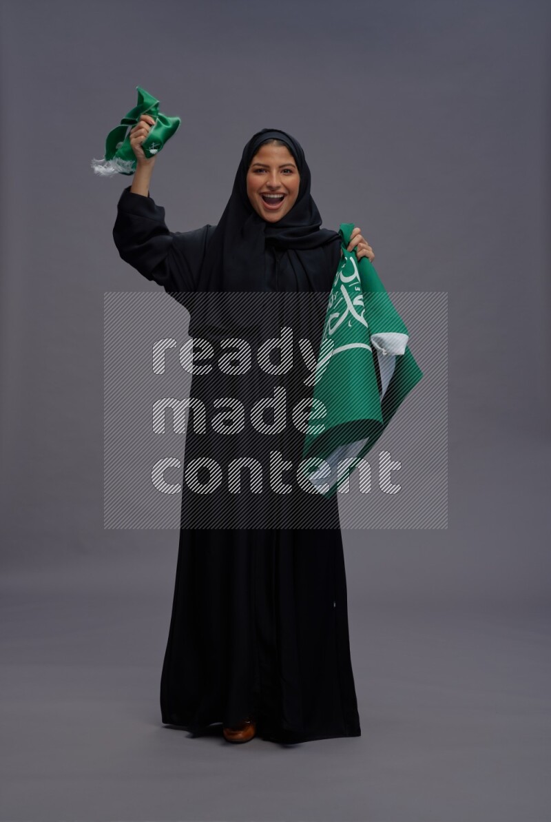 Saudi woman wearing Abaya standing holding Saudi flag on gray background