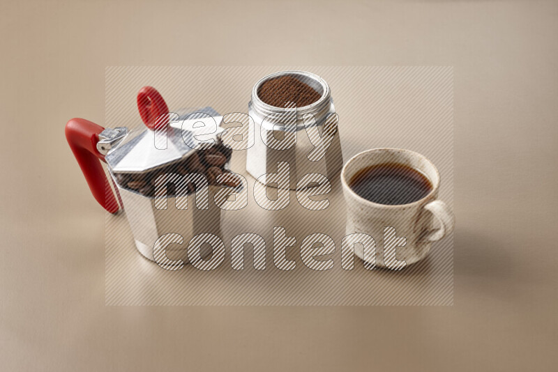 A moka pot with red handle surrounded by roasted coffee beans on beige background