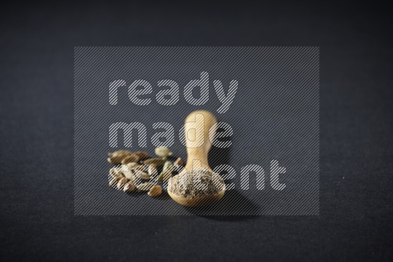 A wooden spoon full of cardamom powder and cardamom seeds beside it on black flooring