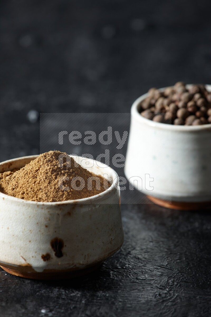 2 beige bowls, one full of allspice powder and the other full of whole balls on a textured black flooring