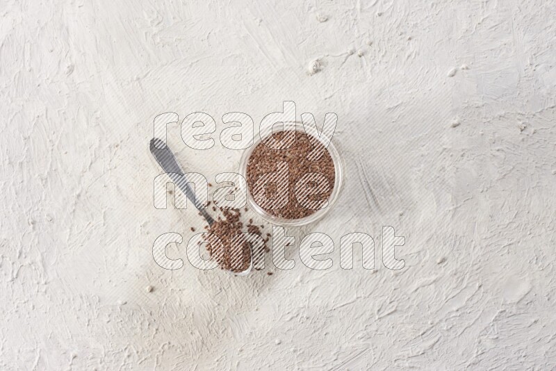 A glass jar full of flax seeds with a metal spoon full of the seeds on a textured white flooring