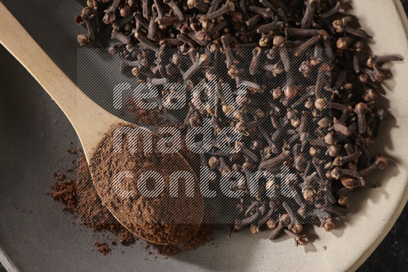 A Pottery plate full of cloves and a wooden spoon full of cloves powder on it on a textured black background
