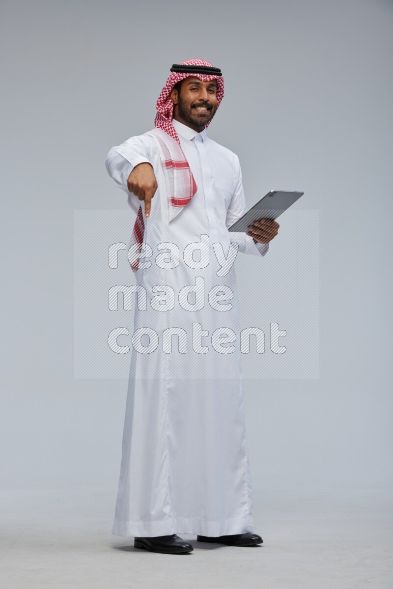 Saudi man Wearing Thob and shomag standing working on tablet on Gray background