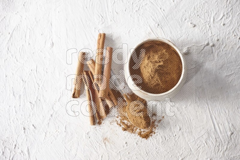 Ceramic beige bowl full of cinnamon powder and a metal spoon with cinnamon sticks next of it on a textured white background