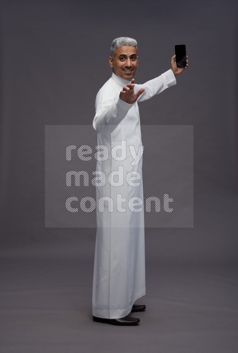 Saudi man wearing thob standing taking selfie on gray background