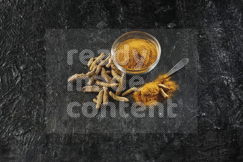 A glass bowl and a metal spoon full of turmeric powder and dried whole fingers next of them on textured black flooring