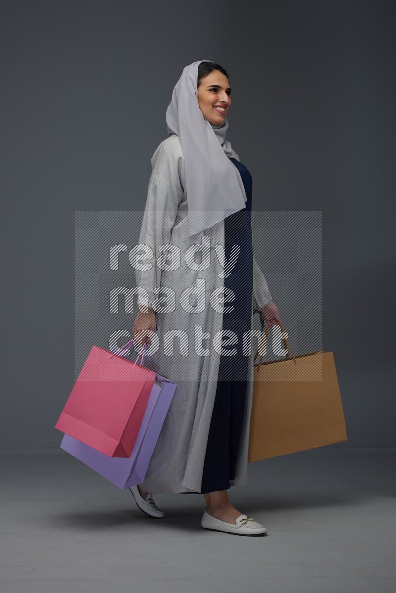 A Saudi woman wearing a light gray Abaya and head scarf standing and holding shopping bags on a grey background