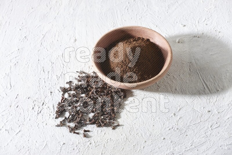 A wooden bowl full of cloves powder with whole cloves beside it on a textured white flooring