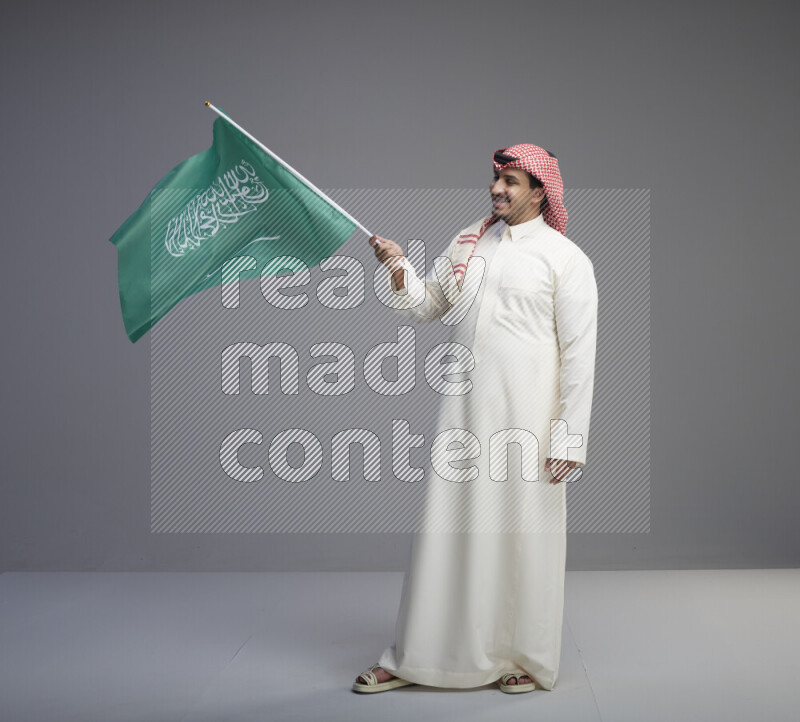 A Saudi man standing wearing thob and red shomag raising big Saudi flag on gray background
