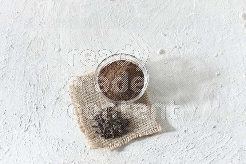 A glass bowl full of cloves powder and cloves grains on a burlap piece on a white flooring