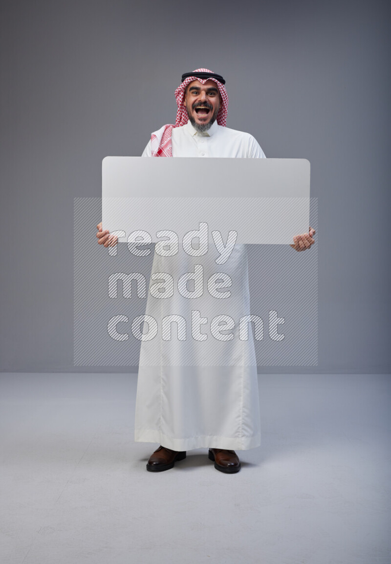 Saudi man Wearing Thob and red Shomag standing holding board on Gray background