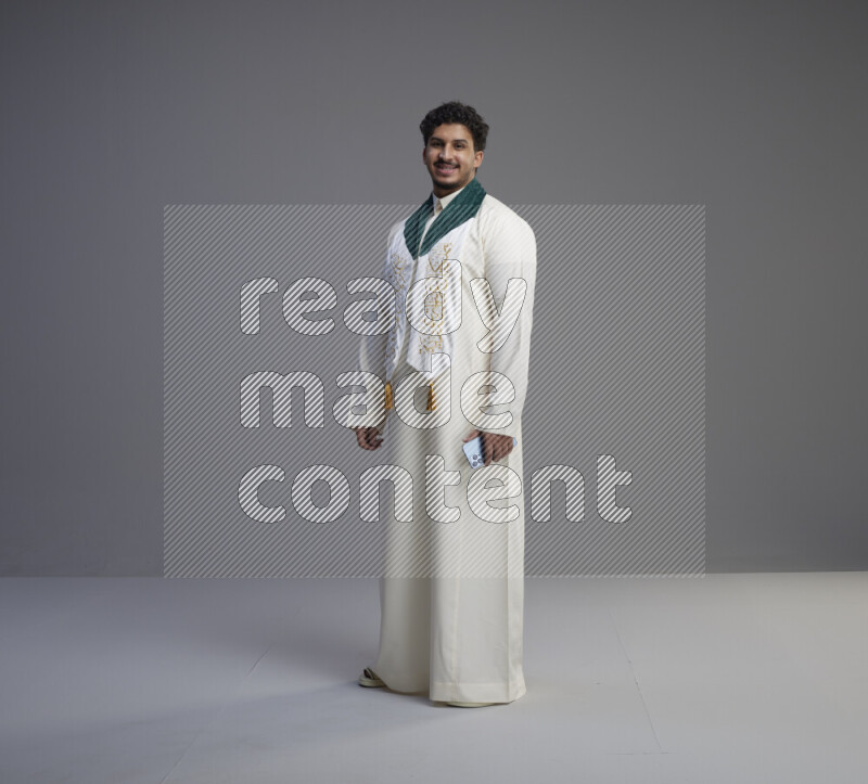 A Saudi man standing wearing thob and Saudi flag scarf on gray background