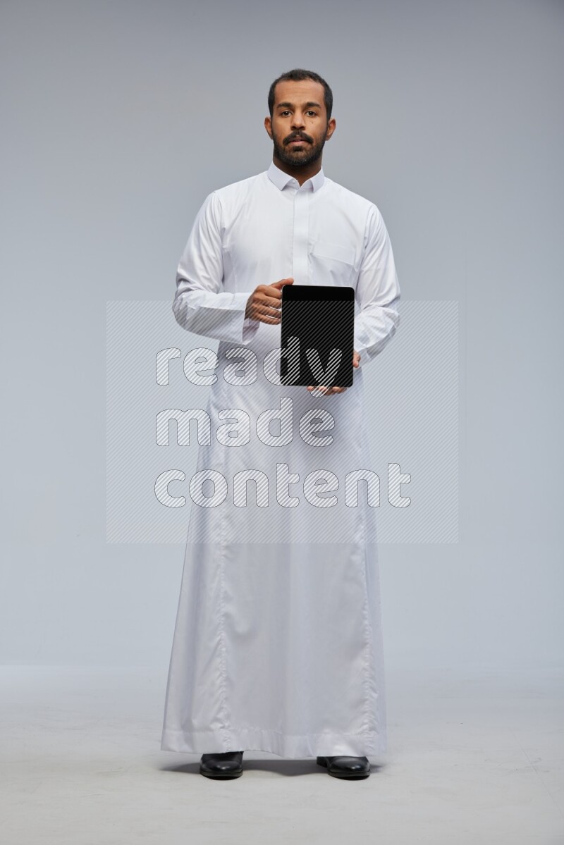 Saudi man wearing Thob standing showing tablet to camera on Gray background