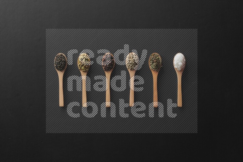 wooden spoons filled with white peppers, cloves, cardamom, salt, black peppers and basil on black flooring