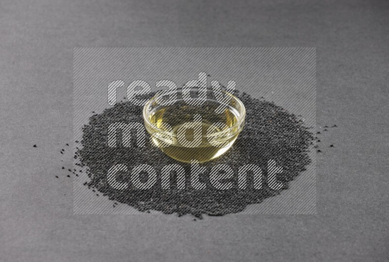 A glass bowl full of black seeds oil surrounded by the seeds on a black flooring